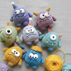 Puede incluir: Una colección de juguetes de monstruos hechos a mano con ganchillo en varios colores, incluyendo morado, verde, amarillo y azul. Cada monstruo tiene características únicas, como cuernos, sombreros y caras expresivas. También se ven ovillos de lana en amarillo, azul y morado.
