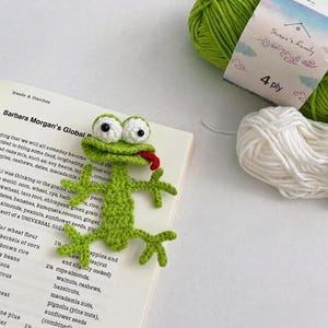 Peut inclure: Un marque-page en forme de grenouille verte au crochet, avec de grands yeux blancs et noirs et une langue rouge. La grenouille est placée sur un livre avec du texte. De la laine verte et blanche est en arrière-plan.
