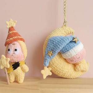 Könnte beinhalten: Zwei handgefertigte Häkelfiguren. Eine ist eine gelbe Figur mit einem Sternenstab und einem rot-orangen Hut. Die andere ist ein schlafendes Baby in einem blau-weißen Hut, das auf einem gelben Halbmond mit goldenen Sternakzenten ruht.