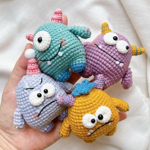 Può includere: Quattro giocattoli mostro all'uncinetto colorati in tonalità di blu, viola e giallo. Ognuno ha un occhio singolo, un sorriso sdentato e braccia piccole. Indossano cappelli da festa e hanno piccole corna. I giocattoli sono fatti a mano e hanno un design stravagante.