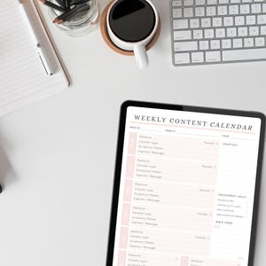 May include: A digital tablet displaying a "Weekly Content Calendar" with a pink and white design. The tablet is on a white surface, alongside a keyboard, coffee cup, notebook, and pencils. The calendar is for planning social media content.
