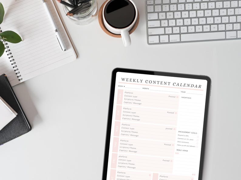 May include: A digital tablet displaying a "Weekly Content Calendar" with a pink and white design. The tablet is on a white surface, alongside a keyboard, coffee cup, notebook, and pencils. The calendar is for planning social media content.
