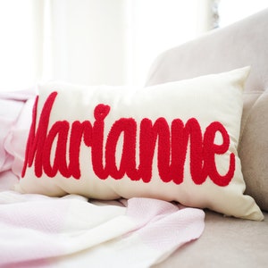 Op de afbeelding: Een wit kussen met de naam "Marianne" geborduurd in rood.