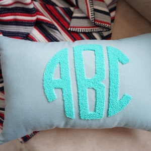 May include: A light blue throw pillow with a turquoise embroidered monogram of the letters "ABC" in a circle.