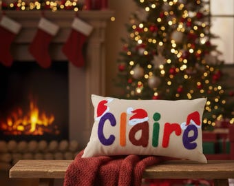 Puede incluir: Un cojín rectangular beige con el nombre "Claire" bordado en letras coloridas, cada una adornada con un gorro de Papá Noel. El cojín descansa sobre un banco de madera cubierto con una manta roja de punto, con un árbol de Navidad y una chimenea al fondo.