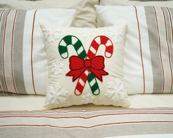 Puede incluir: Una almohada blanca con un diseño de bastón de caramelo rojo y verde y un lazo rojo. La almohada está bordada con copos de nieve.