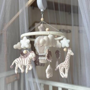 May include: A baby mobile featuring a cream-colored ring with hanging felt shapes. The mobile includes a zebra, giraffe, clouds, stars, and a hot air balloon in neutral tones. The mobile is suspended above a crib.