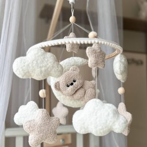 Può includere: Mobile per neonati con un orsetto su una luna, nuvole e stelle. Il mobile è sospeso da una cornice circolare bianca con dettagli in legno, creando una palette di colori morbidi e neutri.