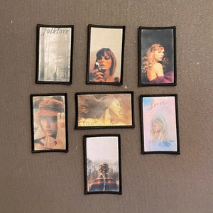 Può includere: Sei patch incorniciati che presentano diverse copertine di album di Taylor Swift. I patch sono incorniciati di nero e presentano immagini di Taylor Swift con i titoli degli album "folklore", "lover" e un patch con una scena di bosco.