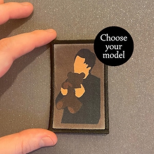 Peut inclure: Un patch brodé marron et noir représentant une silhouette d'une personne tenant un ours en peluche. Le patch a une bordure noire et est sur un fond gris.