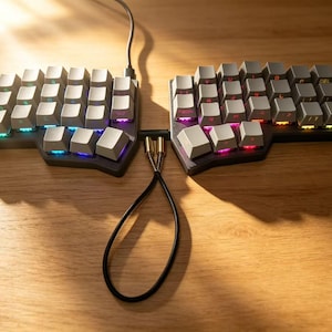 Puede incluir: Teclado ergonómico dividido con teclas grises y retroiluminación LED multicolor. El teclado está conectado por un cable negro con conectores dorados. El teclado está sobre una superficie de madera.