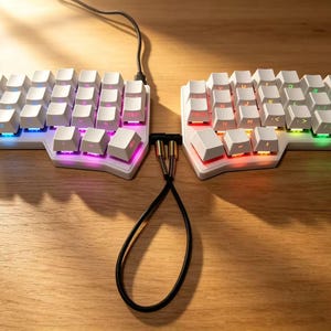 Peut inclure: Un clavier ergonomique divisé avec des touches blanches et un rétroéclairage LED coloré. Le clavier est relié par un câble noir et repose sur une surface en bois. Les touches sont disposées en deux parties.