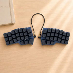 Può includere: Una tastiera ergonomica divisa con tasti blu scuro e una base in similpelle blu abbinata. I tasti sono neri con lettere bianche. Le due metà sono collegate da un cavo nero con connettori color oro. Un taccuino è visibile sullo sfondo.