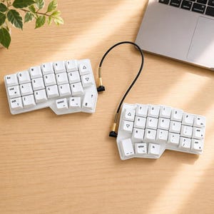 Könnte beinhalten: Zweiteilige weiße ergonomische Tastatur mit schwarzem Verbindungskabel und goldfarbenen Steckern. Die Tastatur hat weiße Tastenkappen mit schwarzer Beschriftung. Die Tastatur liegt auf einer hellbraunen Holzoberfläche.