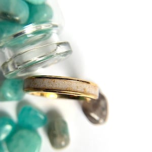 May include: A gold-colored ring with a light-colored band speckled with gold flecks. The ring is set against a white background with scattered turquoise stones and a glass jar.