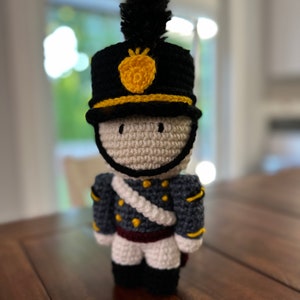 Puede incluir: Un soldado de juguete de crochet que lleva un sombrero negro con un emblema de piña amarillo, un uniforme azul y gris y pantalones blancos.