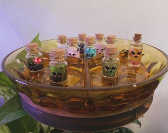 May include: A collection of small glass bottles with cork stoppers, each containing different decorative elements. Some bottles have painted faces, while others hold colorful beads or miniature plants. The bottles are arranged in a divided amber-colored dish.