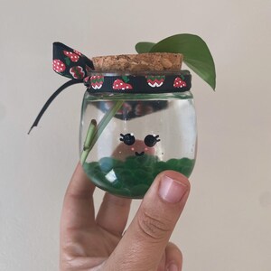 May include: A small, clear glass jar with a cork lid, decorated with a black ribbon featuring a strawberry pattern. The jar contains green beads and a small plant. The jar has a painted face. A hand is holding the jar.