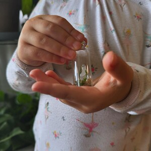 May include: A small glass vial containing a miniature painted ghost figure and greenery. The vial has a silver-colored cap. The ghost is white with black eyes. The background is blurred, suggesting a soft focus.