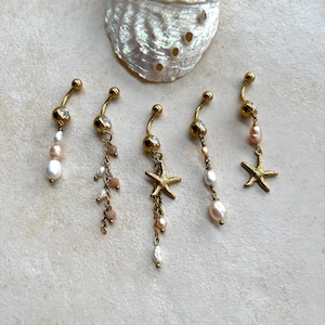 May include: Five gold-toned navel rings featuring clear crystals, small white and pink pearls, and gold starfish charms. Each piece has a curved barbell design with a top ball and a dangling element, arranged below a large iridescent seashell.
