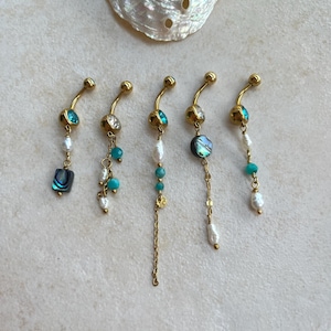 Puede incluir: Cinco piercings dorados para el ombligo con varios dijes colgantes. Los dijes incluyen cuentas turquesas, perlas y detalles de concha de abulón. Los anillos están dispuestos en una fila sobre un fondo neutro, con una concha en la parte superior.