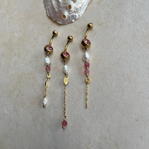 Könnte beinhalten: Drei goldfarbene, gebogene Bauchnabelpiercings mit runden rosa Steinen, weißen Süßwasserperlen und kleinen goldenen Anhängern. Jedes Schmuckstück verfügt über eine zarte Goldkette, wobei eines einen strukturierten, blattförmigen Anhänger besitzt.