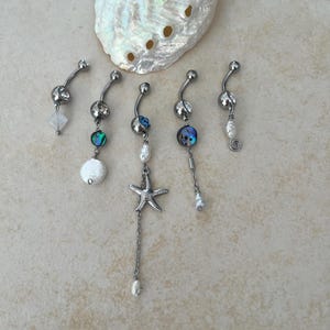 May include: Five silver-toned curved barbell navel rings featuring various dangling charms including a starfish, iridescent abalone shell beads, white pearls, and clear crystals, arranged in a row below a large iridescent abalone seashell.