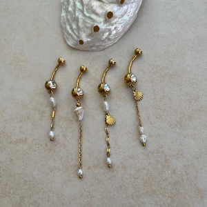 May include: Four gold-toned curved barbell body jewelry pieces with clear round crystals. Each features dangling elements including small white pearls, delicate gold chains, and miniature seashell charms, arranged near a large iridescent abalone shell.