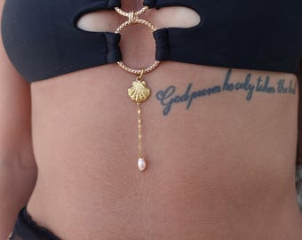 May include: A black bikini top featuring two gold-toned textured circular rings at the center. A gold-toned shell charm hangs from the bottom ring, attached to a delicate gold chain with a small, pale pink teardrop-shaped pearl at the end.