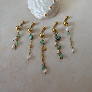May include: Five gold-toned navel rings featuring dangling chains, small white pearls, turquoise beads, and iridescent shell accents. Each piece includes a clear crystal set in the top ball, with varying arrangements of charms and beads.