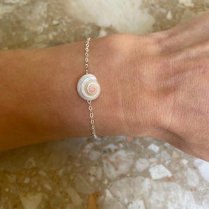 Puede incluir: Una delicada pulsera de plata con un colgante de concha natural. La concha es blanca y beige con un diseño en espiral. La pulsera es de estilo cadena simple, perfecta para el uso diario. La pulsera se muestra en una muñeca.