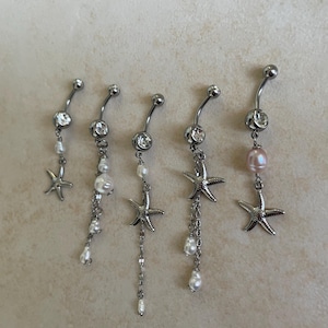 May include: Five silver-toned curved barbell belly button rings featuring clear crystals, dangling chains, small white pearls, and metallic starfish charms. One ring includes a single larger pink pearl accent, while the others feature various arrangements of white pearls and silver chains.