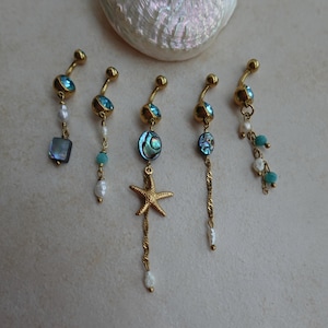 May include: Five gold-toned navel rings featuring blue crystals, white pearls, turquoise beads, and abalone shell accents. One ring includes a gold starfish charm, while others feature dangling chains and various bead combinations.