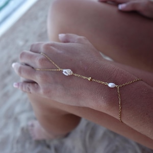 May include: A gold-colored hand chain featuring a seashell and a pearl. The delicate chain connects to a ring and wraps around the hand. The jewelry is worn on a hand with a beach background.