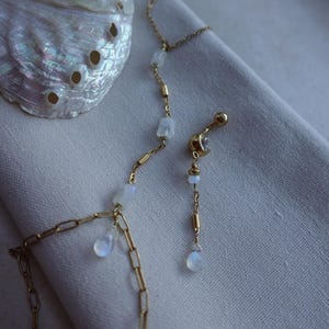 May include: A gold-toned chain necklace featuring small, translucent, iridescent moonstone-like beads and a matching gold-toned navel ring with a single clear stone and a small dangling bead, both showcasing delicate metal link construction.