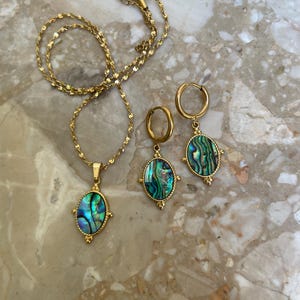 May include: A gold-toned jewelry set consisting of a necklace and a pair of hoop earrings. Each piece features an oval-shaped pendant with an iridescent abalone shell inlay, framed by a textured gold border with small decorative metal accents.