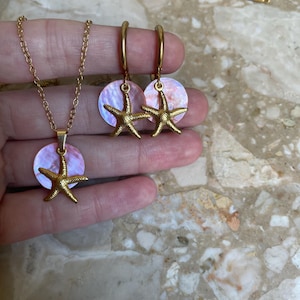 May include: A gold-toned necklace and a pair of matching hoop earrings, each featuring a circular iridescent shell pendant topped with a textured gold-toned starfish charm. The jewelry set includes a delicate gold-toned chain for the necklace.