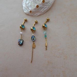 May include: Three gold-tone curved barbell belly rings featuring blue crystals, small white beads, and dangling accents including a square abalone shell charm, a small oval abalone shell, a gold sunburst charm, and a delicate gold chain link.