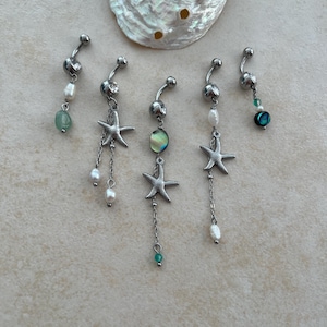 May include: Five silver-toned navel rings featuring dangling charms including metal starfish, white freshwater pearls, and iridescent abalone shell beads. Each piece is constructed with a curved barbell and a top ball, arranged near a large abalone shell.