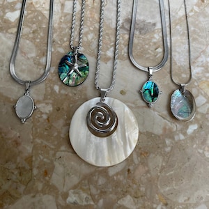 May include: A selection of silver necklaces with different pendants. The pendants feature an oval mother-of-pearl, a starfish on an abalone shell, a large spiral design, and an abalone shell. The necklaces are presented on a marble surface.