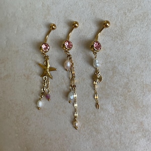 Può includere: Tre piercing per ombelico color oro con pietre preziose rosa e ciondoli decorativi. Uno presenta una stella marina, un altro perle pendenti e il terzo un sole con perle. I gioielli sono esposti su uno sfondo neutro.