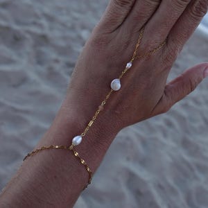 May include: A gold-colored hand chain with a delicate design. The chain features several white pearls and small, gold-colored beads. The chain connects to a bracelet and extends up the hand to a ring.
