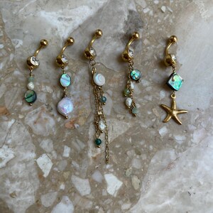 May include: Five gold-colored belly button rings with various dangling charms. Charms include abalone shells, pearls, clear crystals, and a gold starfish. The rings are displayed on a light-colored, speckled surface.