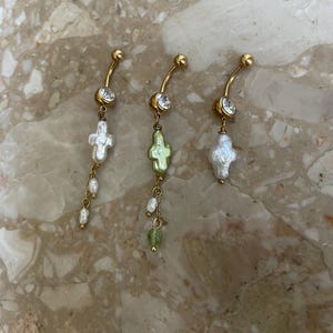 Könnte beinhalten: Drei goldfarbene Bauchnabelpiercings. Jeder Ring hat ein gebogenes Stangendesign mit einem klaren Edelstein oben. Ein Ring hat einen weißen kreuzförmigen Anhänger, ein anderer einen grünen kreuzförmigen Anhänger und der dritte einen weißen kreuzförmigen Anhänger.