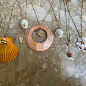 Peut inclure: Cinq colliers en or avec différents pendentifs en forme de coquillages et d'étoiles de mer. Les colliers sont posés sur une surface texturée brun clair.