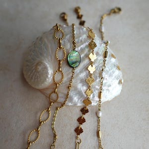 Può includere: Collane dorate con vari design, tra cui maglie ovali, un ciondolo di abalone verde e ciondoli a forma di conchiglia. Le collane sono esposte su una conchiglia bianca, creando un'estetica da spiaggia. I gioielli sono delicati ed eleganti.