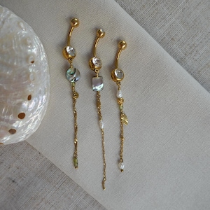 May include: Three gold-toned curved barbell navel rings featuring dangling chains. Each piece includes a clear round stone, a metallic bead, and accents of iridescent shell or small white pearls. The jewelry is displayed alongside a large white seashell.