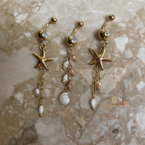 May include: Three gold-colored belly button rings with a beach theme. Each ring has a curved barbell design with a clear gem at the top. One ring features a starfish charm, while the others have pearl and shell accents.