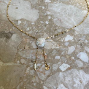 May include: A gold-coloured necklace with a spiral seashell pendant. The necklace has a fine chain with small gold beads and two dangling charms: a pearl and a seashell. The background is a marble surface.