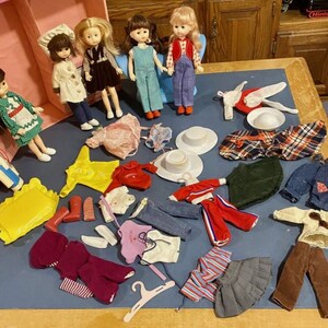 Può includere: Una collezione di bambole vintage e i loro vestiti. Le bambole sono vestite con abiti diversi, tra cui un vestito a quadri rosso e bianco, un vestito in denim blu e un vestito rosa e bianco. I vestiti includono un impermeabile giallo, una gonna a righe rosso e bianco e una gonna in denim blu.